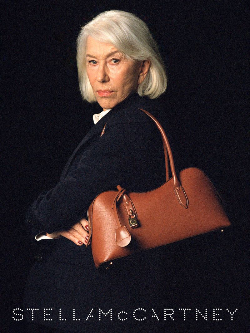 Helen Mirren wearing a dark blazer holding a brown leather shoulder bag against a black background for Stella McCartney