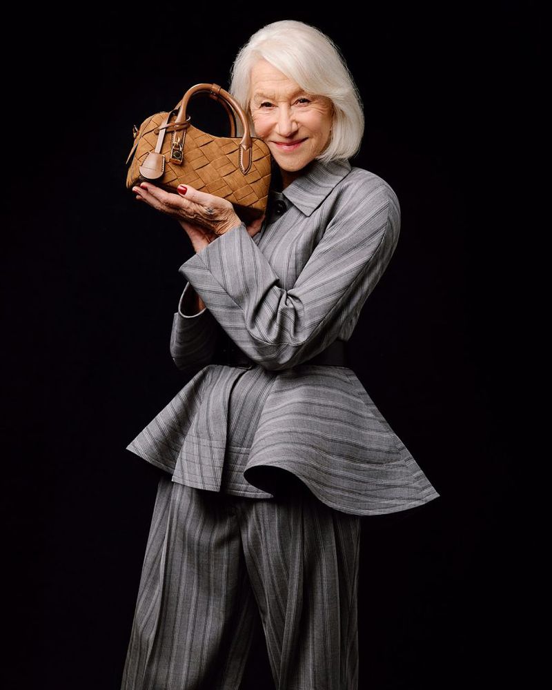 Helen Mirren wearing a grey pinstripe suit holding a brown woven handbag against a dark background for Stella McCartney