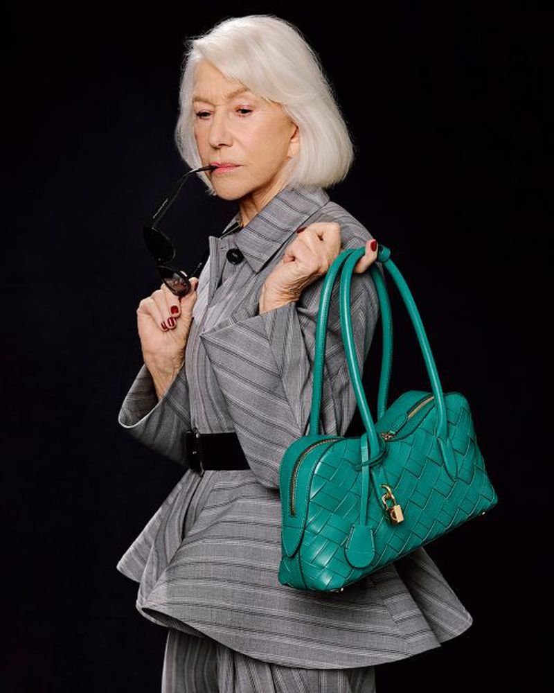 Helen Mirren wearing a grey pinstripe jacket and holding a teal leather handbag against a dark background