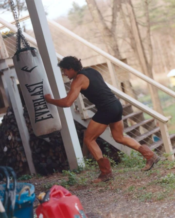 Man wearing a dark tank top and brown cowboy boots punching a heavy punching bag outdoors near a wooden porch