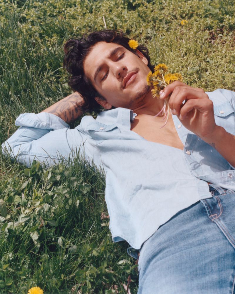 Man laying on a grassy field holding yellow wildflowers with a relaxed facial expression, wearing an unbuttoned light blue shirt and blue jeans.