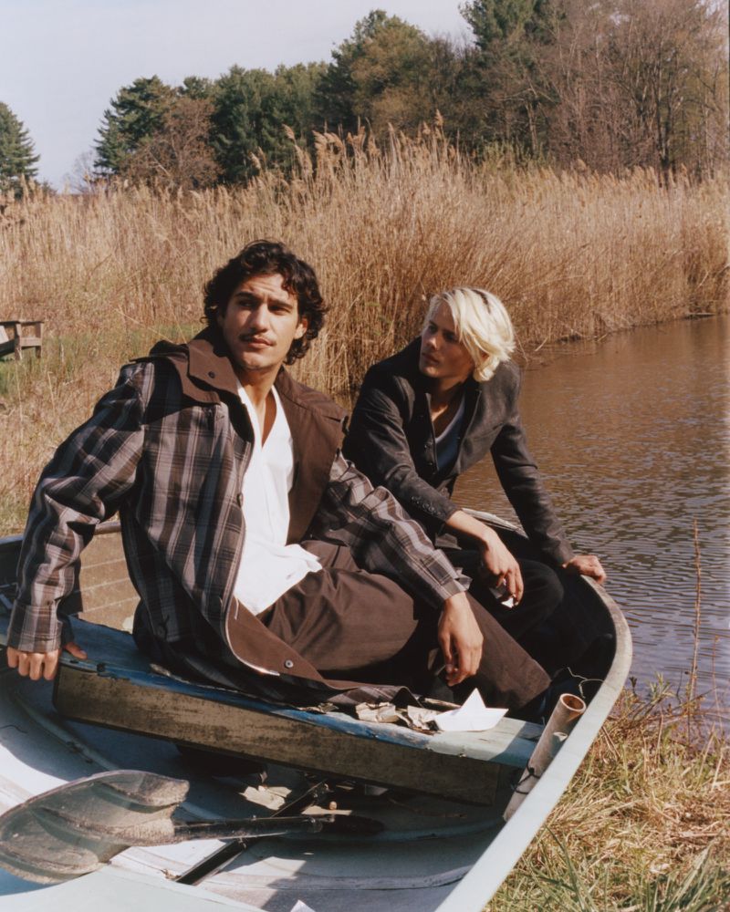 Two models seated inside a small rowing boat near a grassy riverbank outdoors.