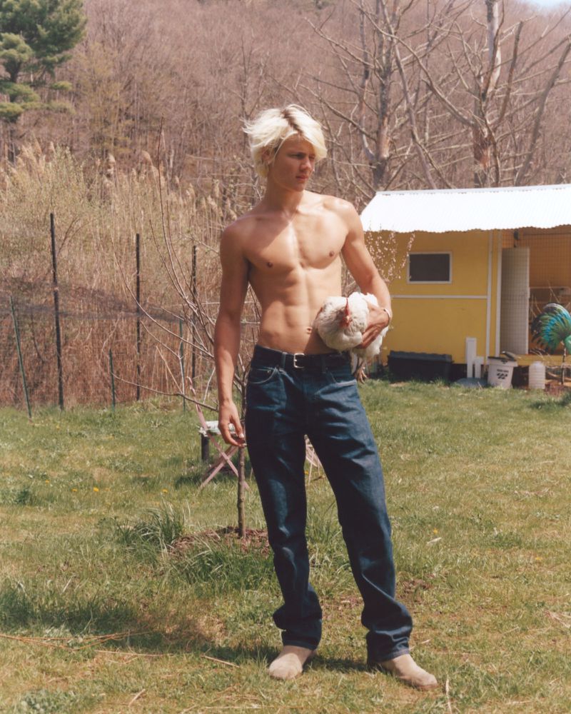 Shirtless young man with blonde hair wearing blue jeans holding a white chicken outdoors
