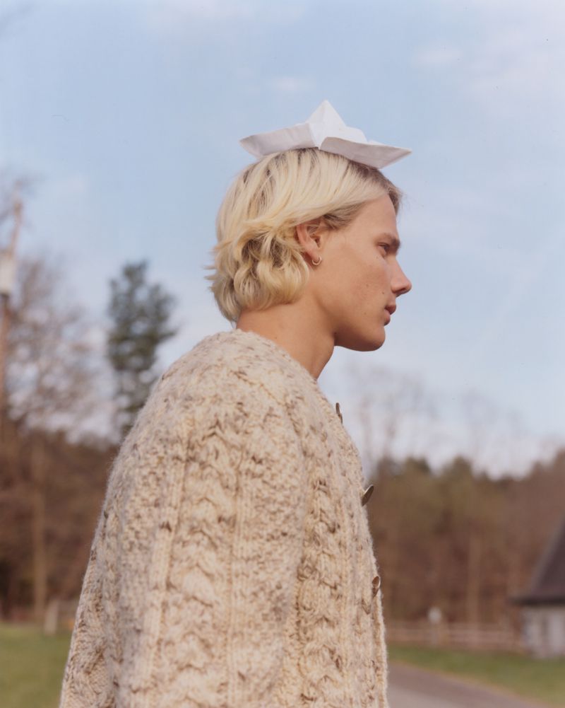 Profile of a blonde man wearing a paper hat and textured knit cardigan outdoors under an overcast sky.