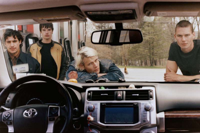 Four men looking through the windshield of a car from outside, shot from the driver seat interior.