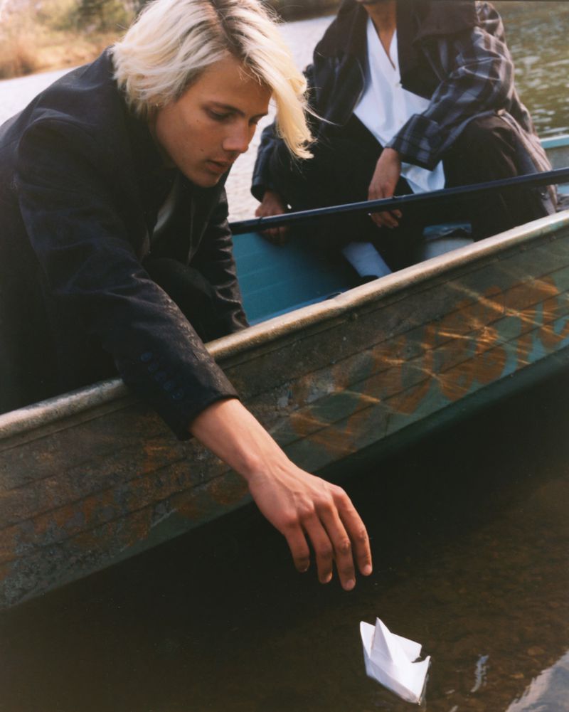 Young man with blonde hair leaning from a canoe to touch a small paper boat floating on the water.