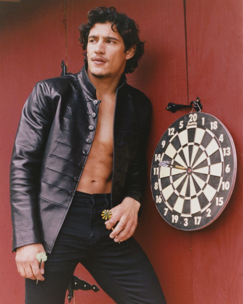 Man wearing a black leather jacket and black trousers leaning against a red wall next to a dartboard