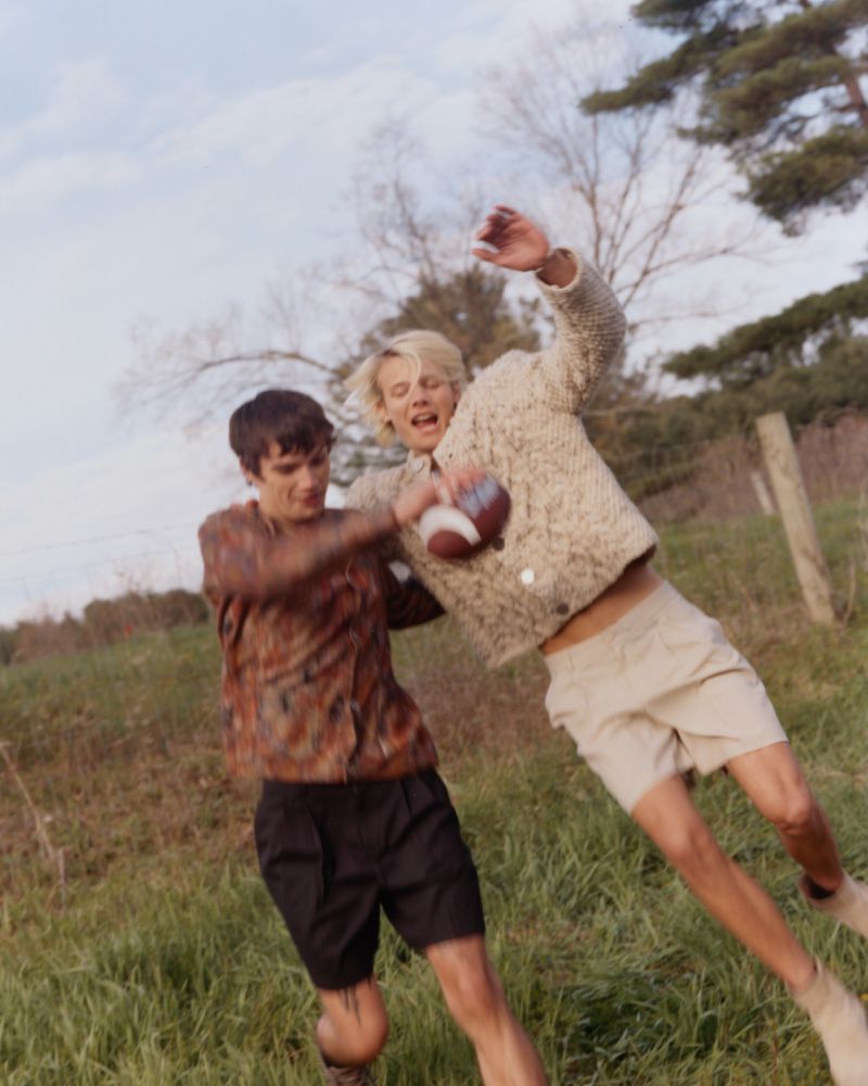 Two young men in motion outdoors in a grassy field, one jumping with a football while the other reaches for it.