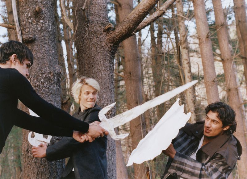Three young men play-fighting with white paper swords in a wooded forest setting.