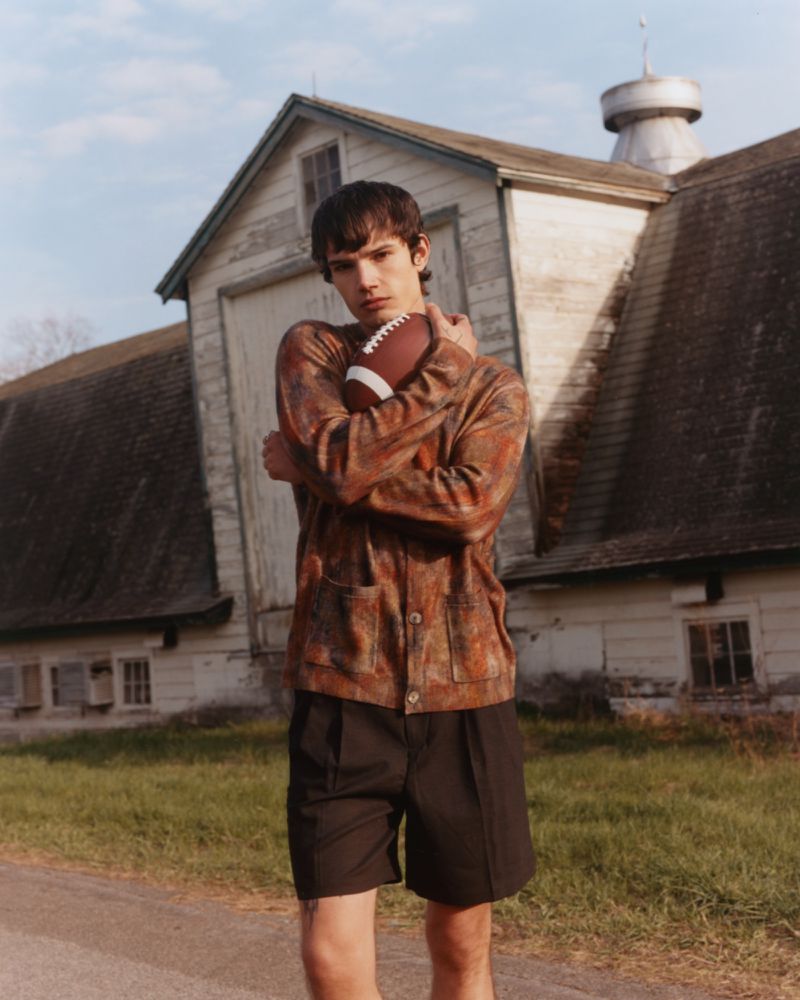 A young male model holding a football in front of a rustic wooden barn structure for a Numéro New York fashion editorial.