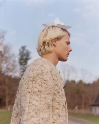 Profile of a blonde man wearing a paper hat and textured knit cardigan outdoors under an overcast sky.