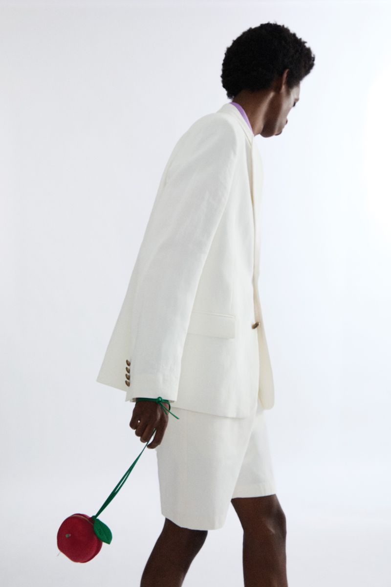 Model in a white suit jacket and shorts holding a red fruit-shaped bag against a seamless white background.