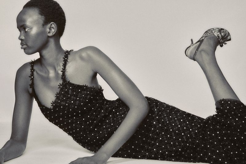 Black and white fashion portrait of a model lying down wearing a dark floral embellished dress against a seamless white studio background.