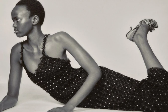 Black and white fashion portrait of a model lying down wearing a dark floral embellished dress against a seamless white studio background.