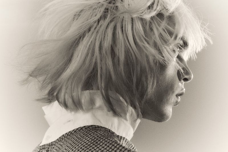 Profile portrait of a model with voluminous blonde hair and patterned clothing against a plain background.