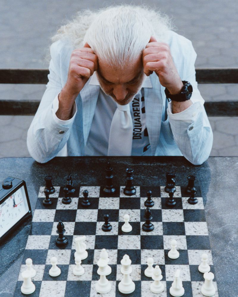 An older man with long white hair holding his head while playing a game of chess outdoors.