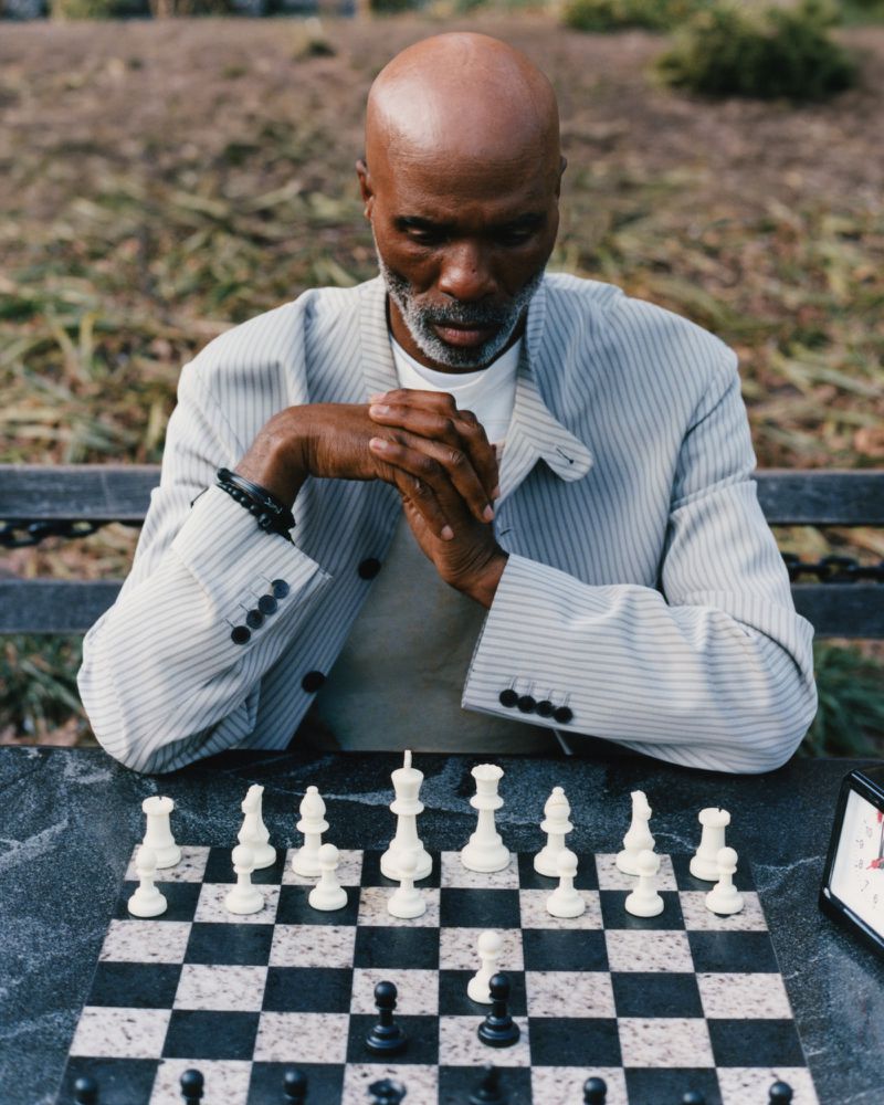 Black man with gray beard seated outdoors playing chess, wearing a light gray striped jacket.