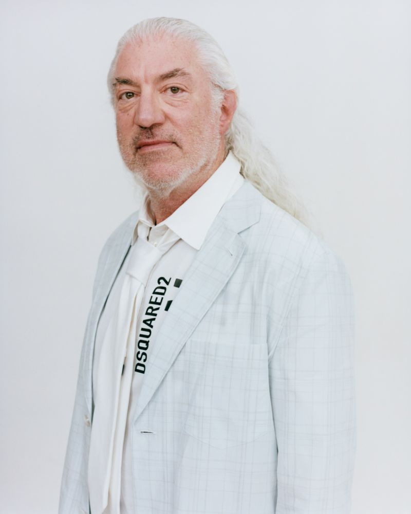 Portrait of an elderly man with long white hair and beard wearing a light blazer and white tie against a plain white background