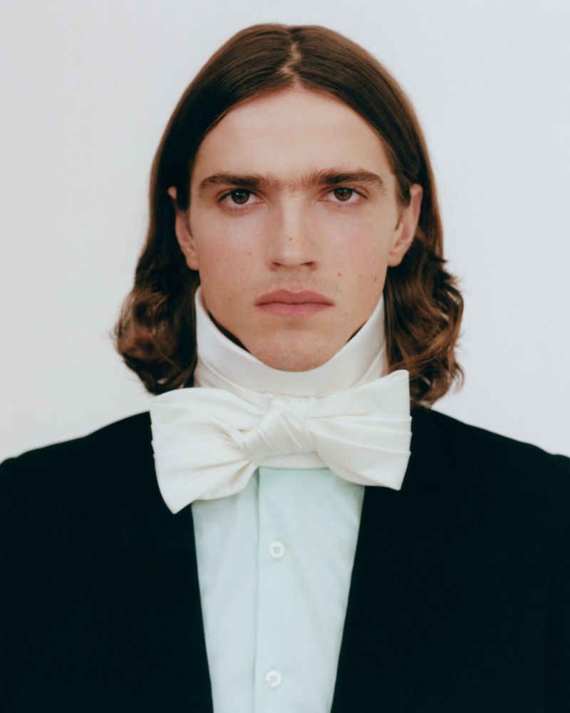 Portrait of a man with long brown hair wearing a black jacket and a large white bow tie against a white background