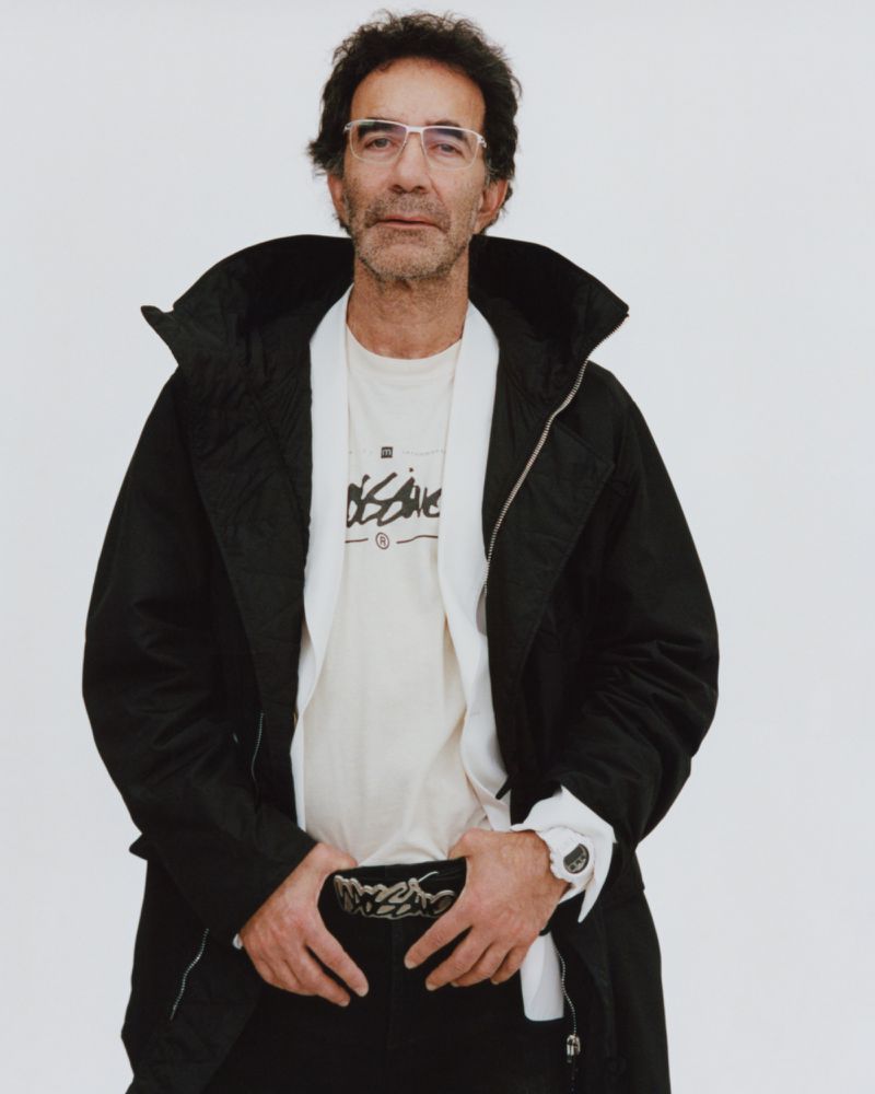 Middle-aged man with glasses in dark jacket over graphic tee, hands near belt, posing against a plain white background in a medium studio shot.