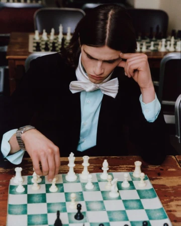 Young man with long dark hair playing chess, wearing a black jacket, light blue shirt, and white bow tie