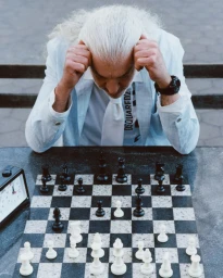 An older man with long white hair holding his head while playing a game of chess outdoors.