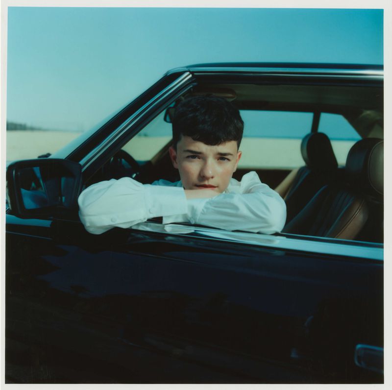 A young man leans against the window of a classic black car parked near a beach with a light blue sky in the background.