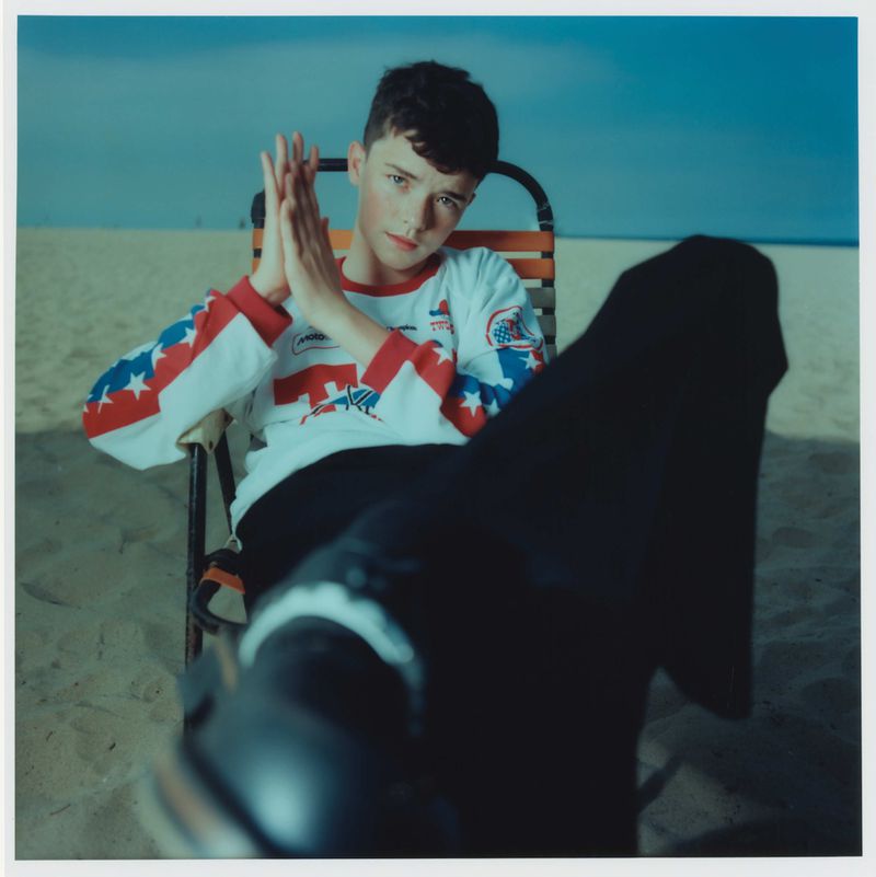 Young adult seated on a beach chair in a red, white, and blue shirt on a sunny beach with blue sky and sand in the background.