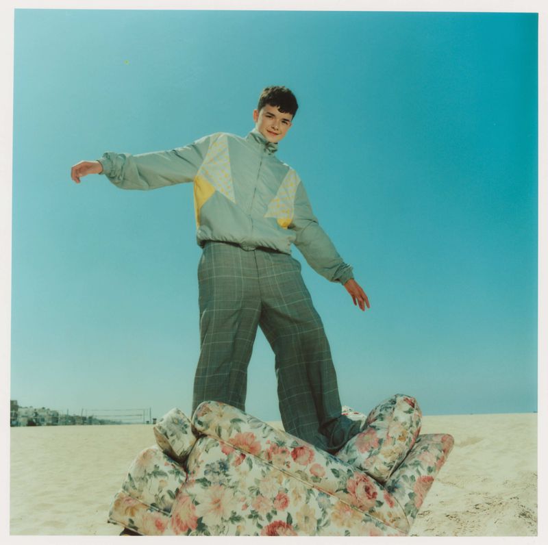 A young man balances on a floral-patterned armchair on a beach under a clear blue sky, wearing a tracksuit with a grey and yellow jacket.