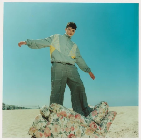A young man balances on a floral-patterned armchair on a beach under a clear blue sky, wearing a tracksuit with a grey and yellow jacket.