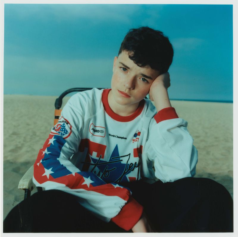 Young person sits on a beach chair in front of a sandy beach and light blue sky in an editorial-style outdoor portrait.