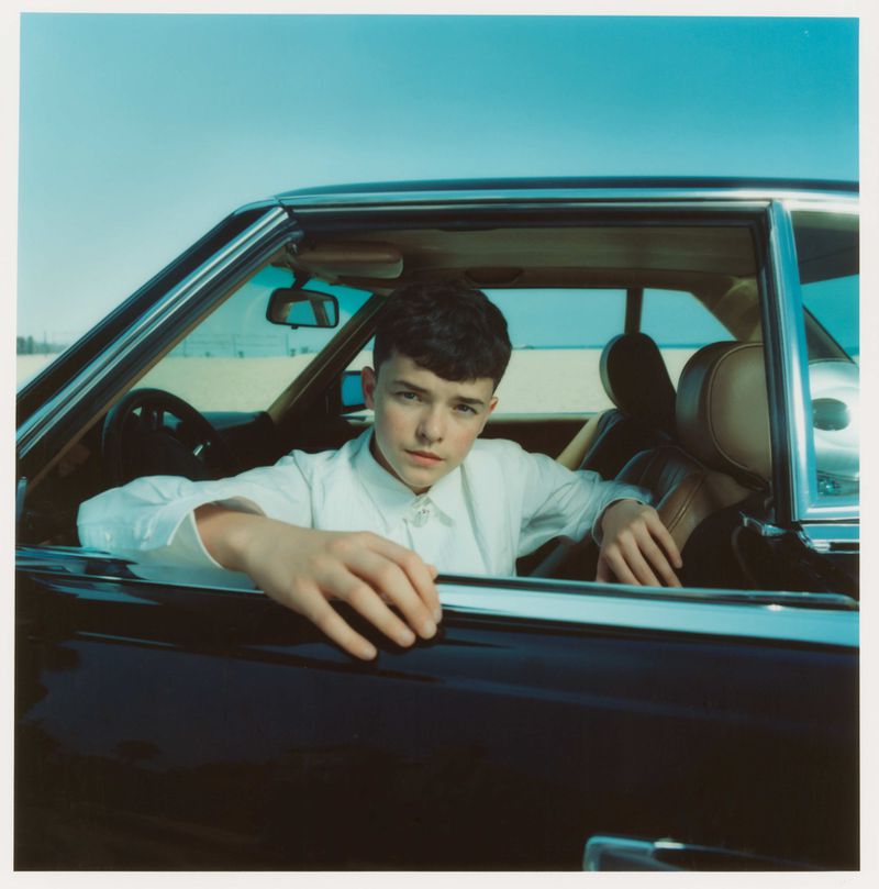 Young adult poses in a classic car on a beach: Close-up portrait capturing a blend of automotive and coastal elements under daylight.