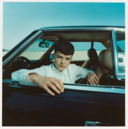 Young adult poses in a classic car on a beach: Close-up portrait capturing a blend of automotive and coastal elements under daylight.