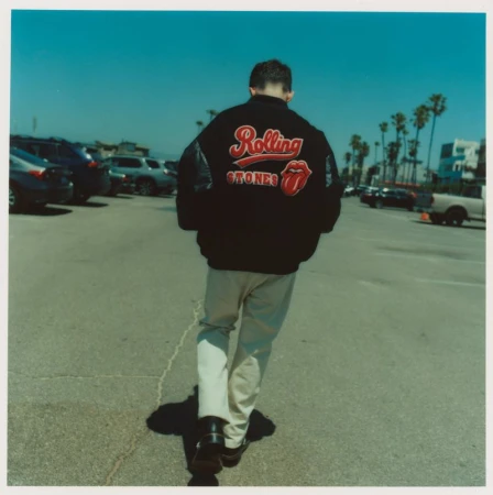 A person walks away in a parking lot wearing a black jacket with 'Rolling Stones' logo against a blue sky with palm trees.