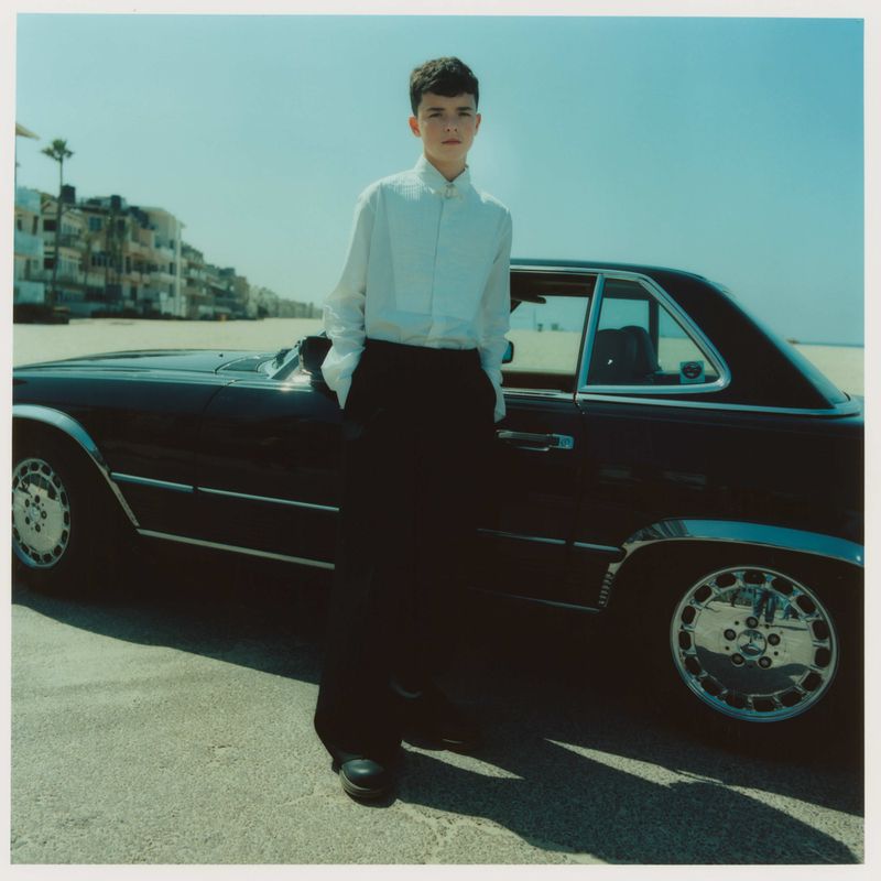 A young adult stands by a black vintage convertible car on a beach road with palm trees and buildings in the background on a sunny day.