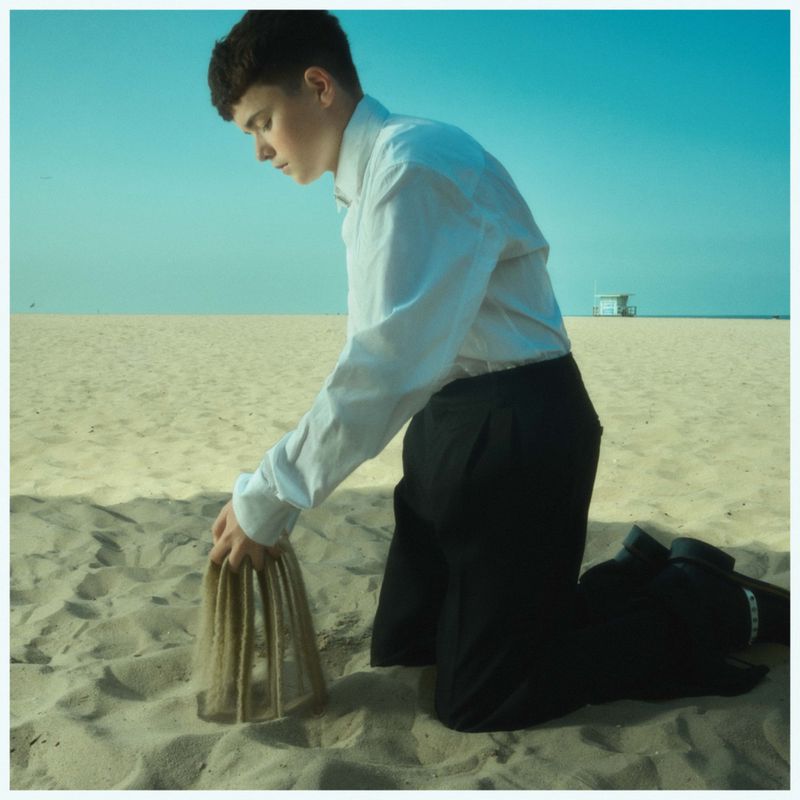 Young adult kneeling on a sandy beach, forming sand sculpture with hands, wearing white shirt and dark pants against a clear sky.