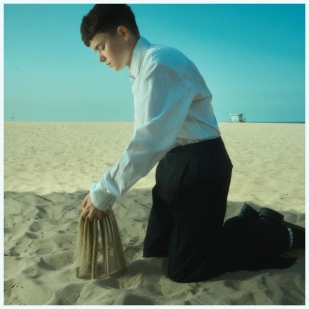 Young adult kneeling on a sandy beach, forming sand sculpture with hands, wearing white shirt and dark pants against a clear sky.