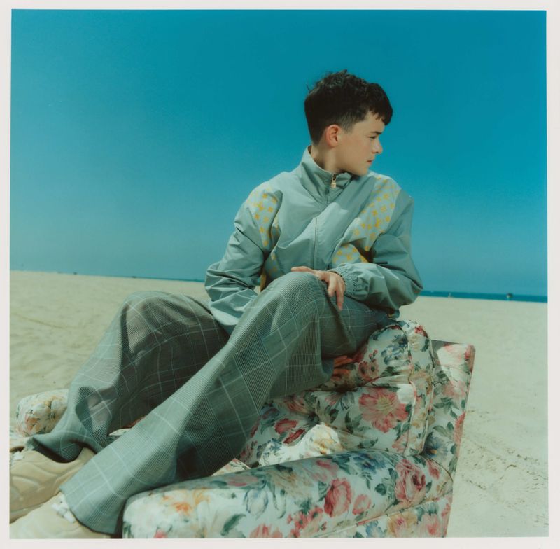 Young adult poses in an armchair on the beach in front of a blue sky. They wear a grey jacket with pattern and checkered pants.