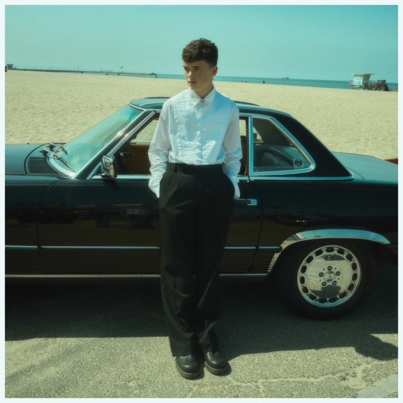 A young man in a white shirt and black pants stands next to a classic black car on a sunny beach with a lifeguard tower in the distance.
