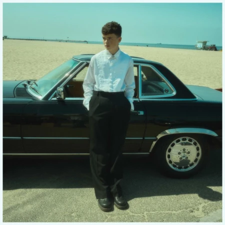 A young man in a white shirt and black pants stands next to a classic black car on a sunny beach with a lifeguard tower in the distance.