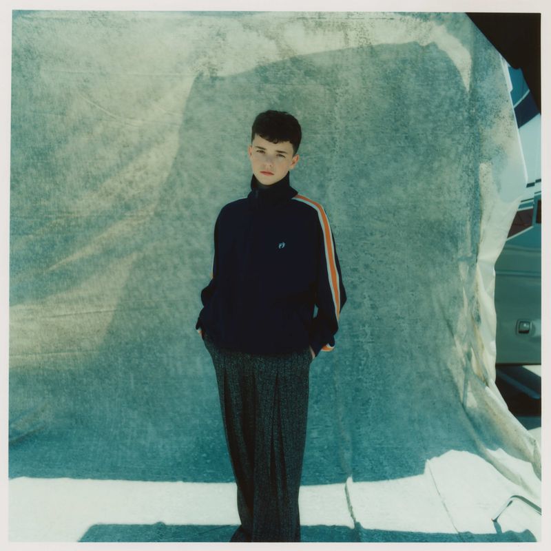Full-length portrait of a young person with short dark hair wearing a navy turtleneck sweater and patterned grey pants against a draped backdrop.