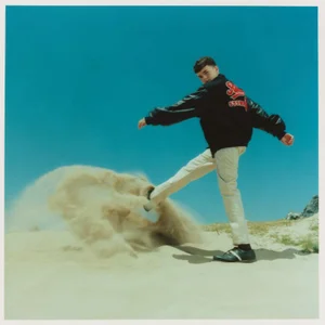 Young man in a black jacket and light pants kicking sand on a beach under a clear blue sky