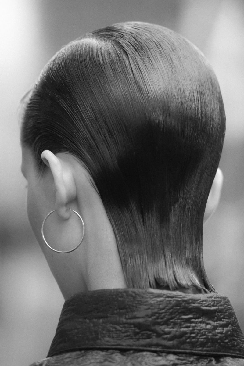 Close-up back view of a woman with slicked-back dark hair wearing a silver hoop earring and a dark textured top.