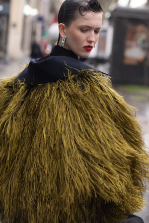 Model in a textured olive feather coat and dark collar with slicked-back hair against an outdoor street background.