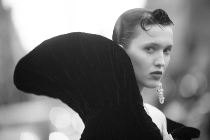 Monochrome portrait of a woman wearing a large sculptural black collar and an ornate drop earring, captured in profile.