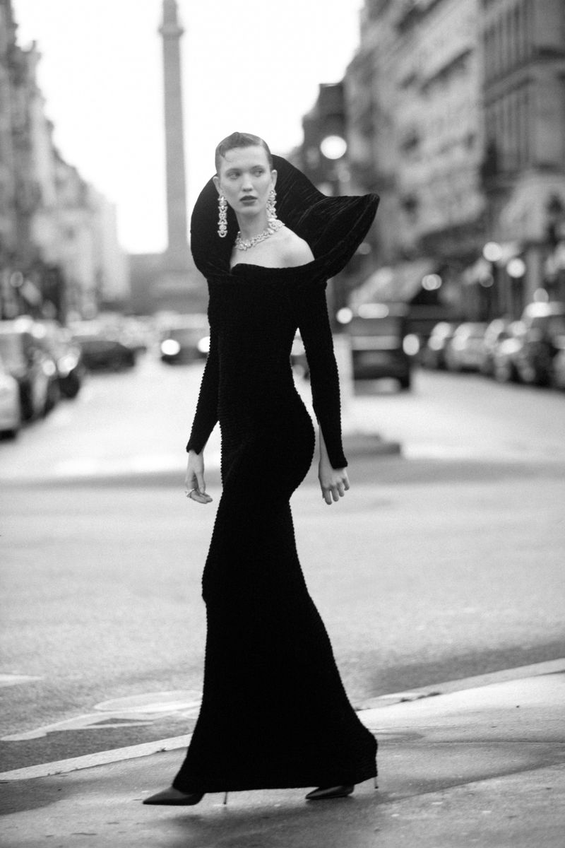 Model in a black evening gown with a sculptural collar walking down a city street in a black and white fashion editorial.