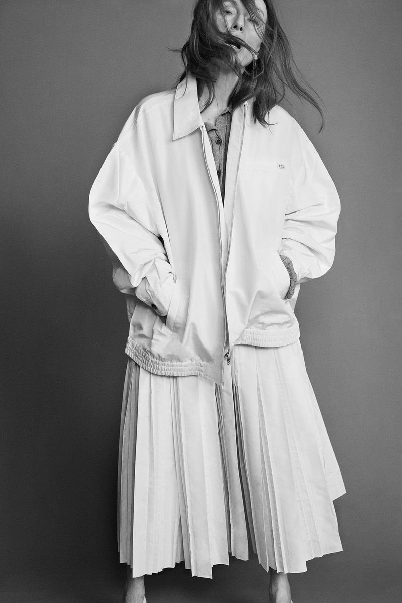 A woman with flowing hair poses in an oversized white jacket and pleated skirt against a grey backdrop in a monochromatic fashion shot.