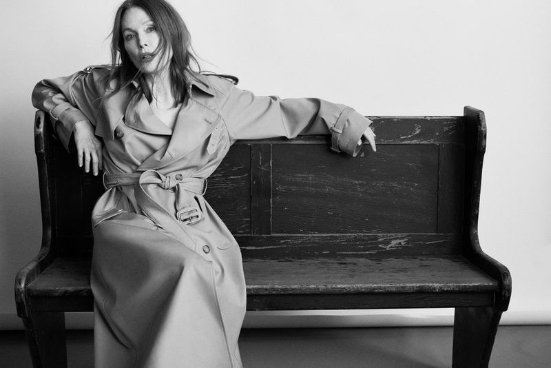 Julianne Moore seated on a dark wooden bench wearing a belted trench coat against a plain light background.