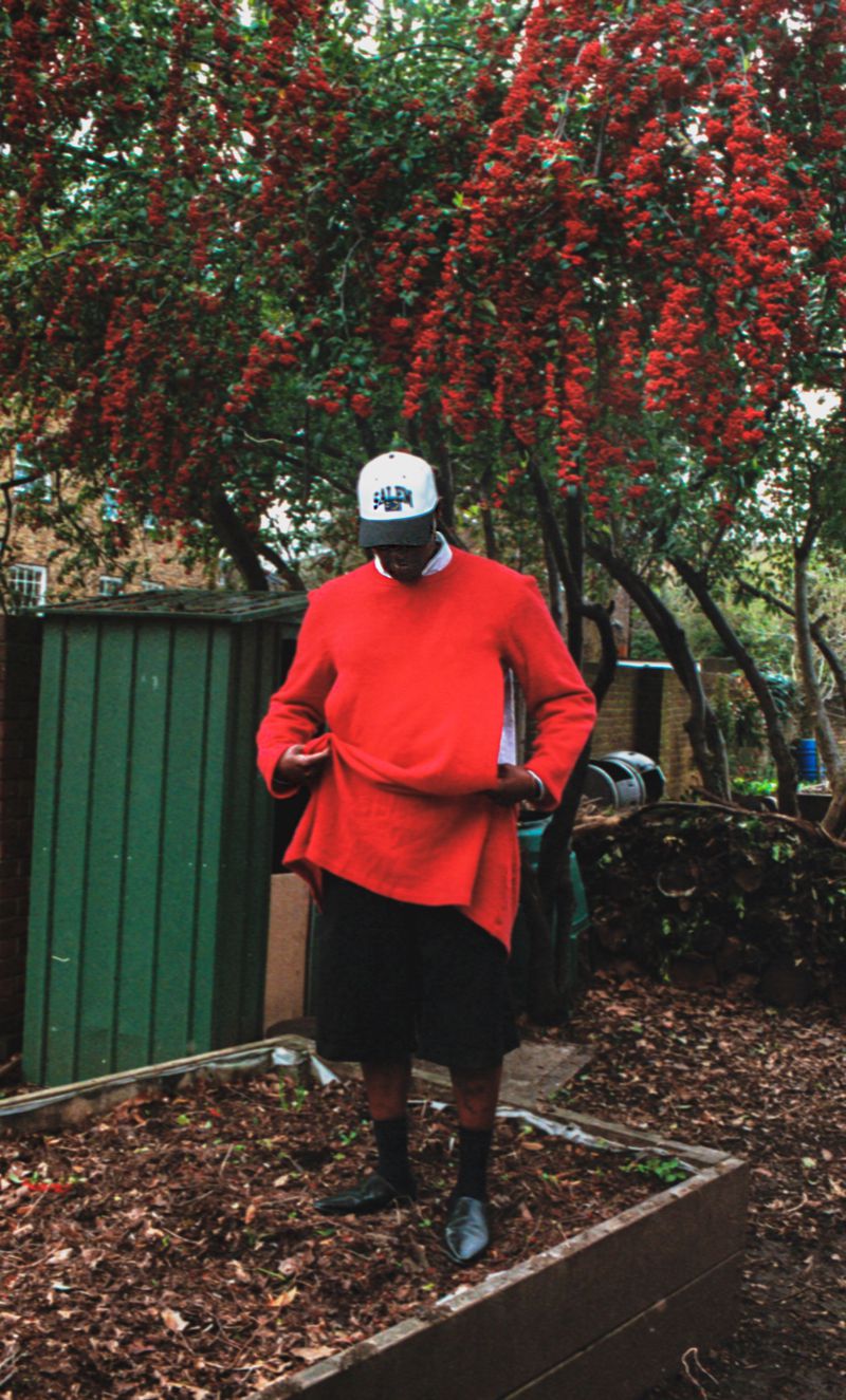 Man wearing a vibrant red sweater and white baseball cap standing outdoors in a garden before a flowering tree for Buffalo Zine.