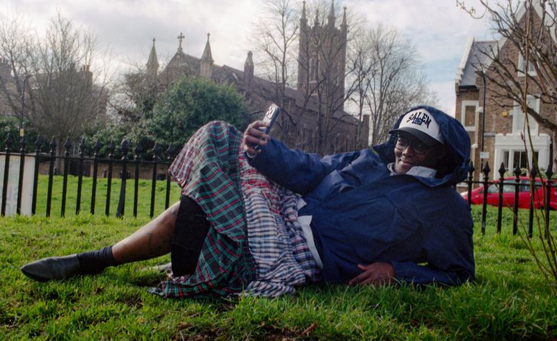 Black man wearing a blue waterproof jacket and plaid skirt reclining on a grassy park lawn, holding up a smartphone.